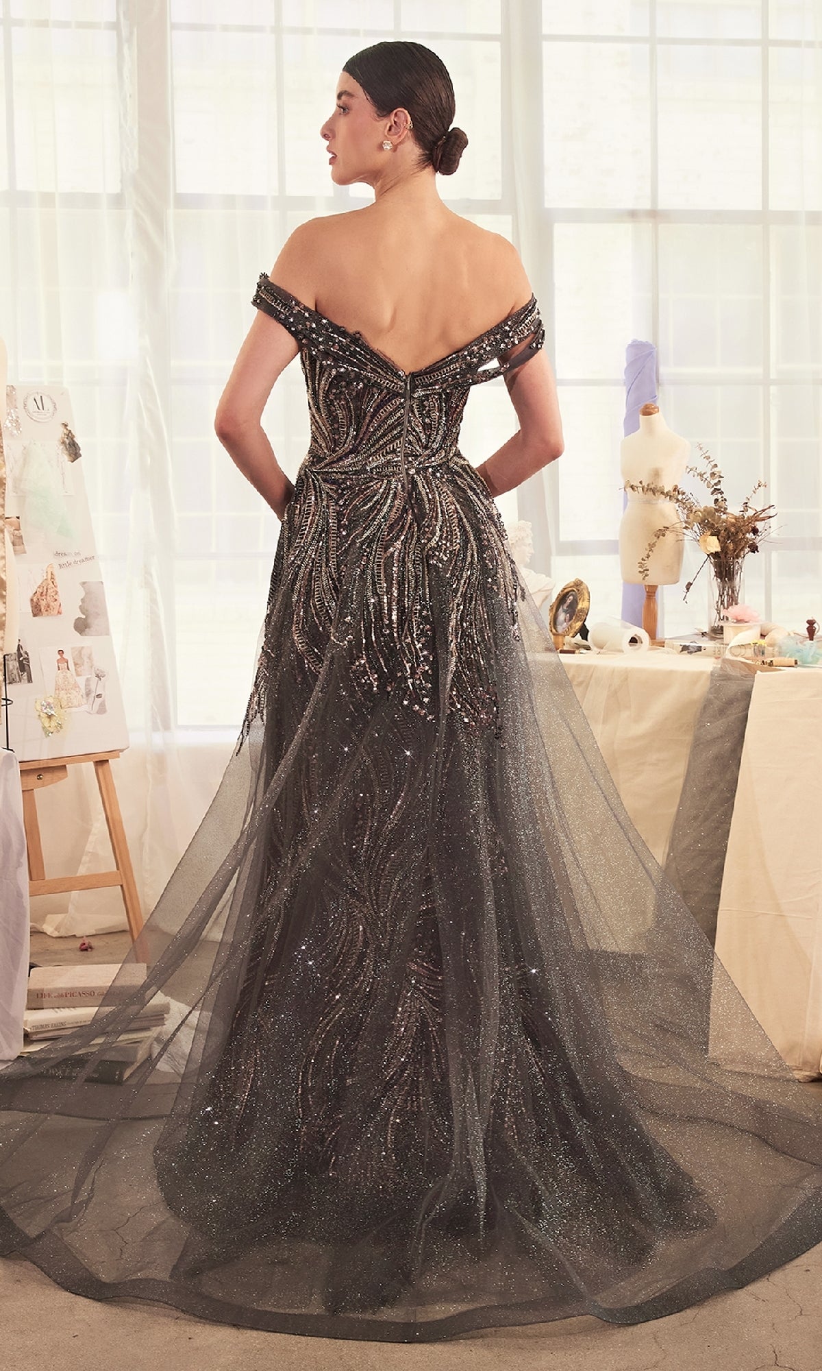 A woman stands in a studio, facing away, wearing the Long Dark Silver Formal Dress: Andrea & Leo A1375 with beaded details and a tulle train. Sketches and a dress form in the background highlight its elegant, classic prom style.